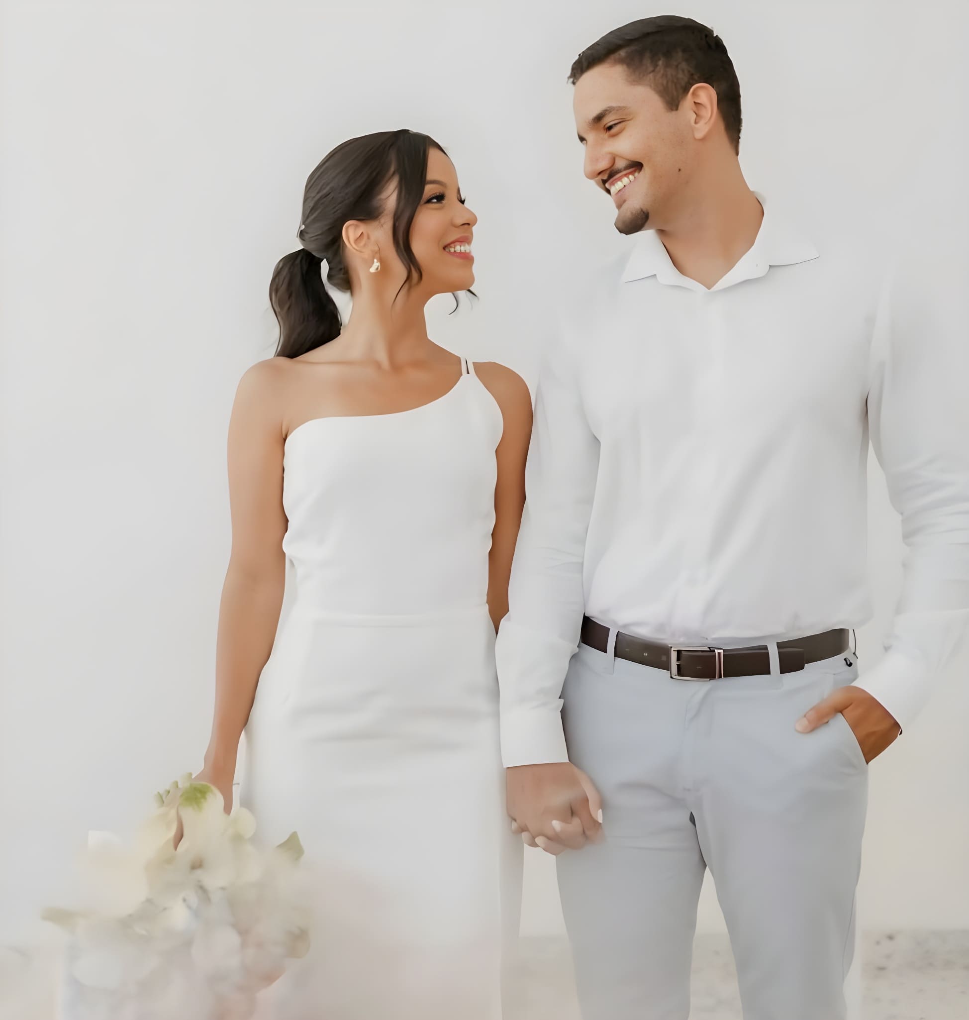 Novios sonriéndose el uno al otro, tomados de la mano, con la novia en vestido blanco y el novio de camisa blanca y pantalón claro. Fotografía de bodas en Uruguay.